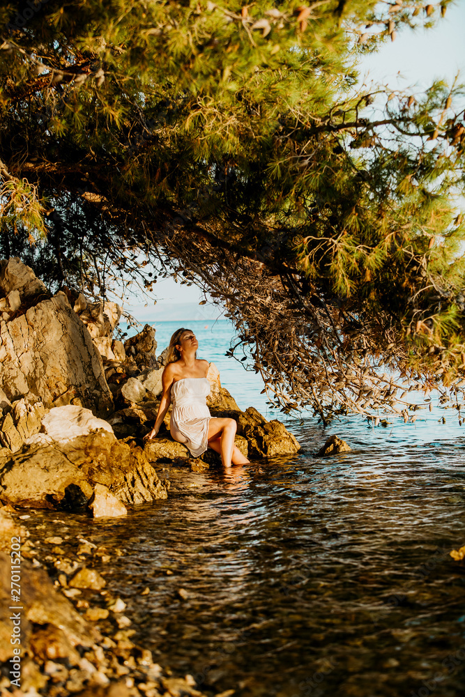 Beautiful young pregnant woman with white wet dress relax on the rocks near the sea water. Summer at sunset. Concept Love and pregnancy