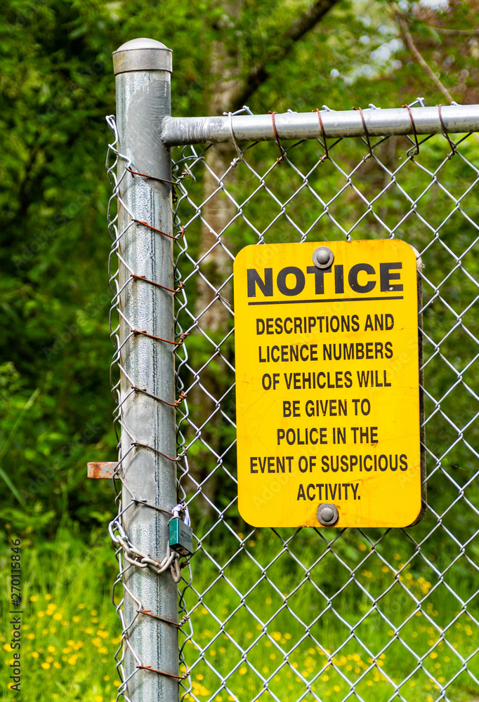 Yellow and black neighborhood crime warning sign posted on chain link fence with wooded background.