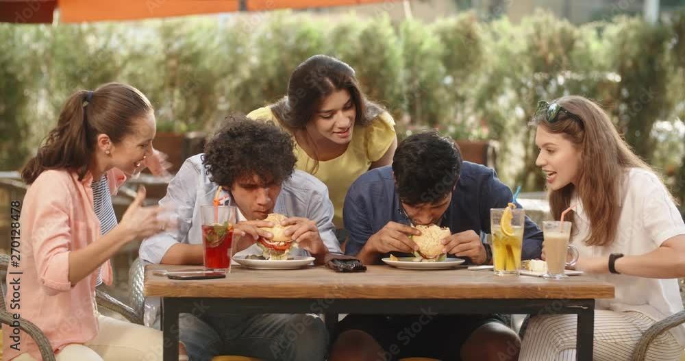 Two male friends having burger-eating battle, girls around cheering and supporting. People enjoying their time in cafe. 4k Two male friends having burger-eating battle, girls around cheering and supporting. People enjoying their time in cafe. 4k