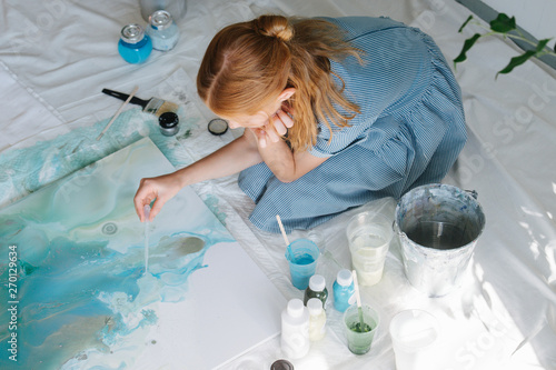 Teenage redhead girl is painting with brush on big canvas on floor in a workshop.