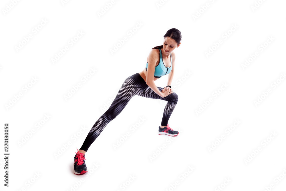 . A young woman in comfortable sportswear (leggings and top) is smiling charmingly and doing wide lunges to the sides with her legs on an isolated white background. . A young woman in comfortable sportswear (leggings and top) is smiling charmingly and doing wide lunges to the sides with her legs on an isolated white background.