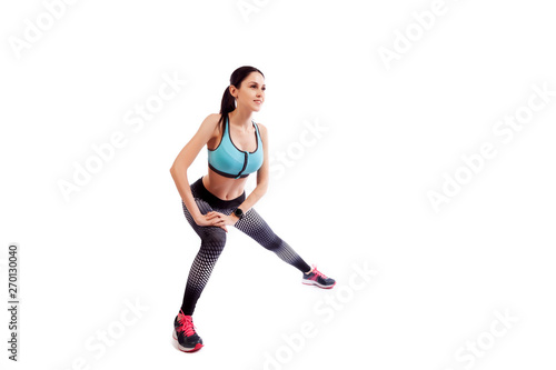  A young woman in comfortable sportswear (leggings and top) is smiling charmingly and doing wide lunges to the sides with her legs on an isolated white background. © Виталий Сова