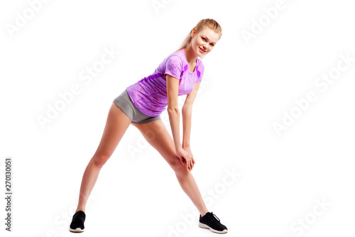  A young woman in comfortable sportswear (shorts and top) is smiling charmingly and doing wide lunges to the sides with her legs on an isolated white background.