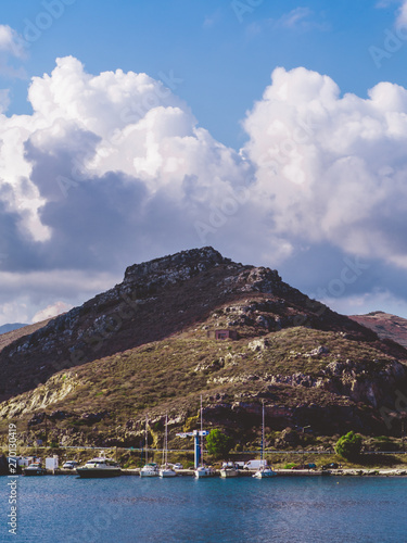Ships docked in a small port beneath the small hill with huge clouds looming over © Dimitrius