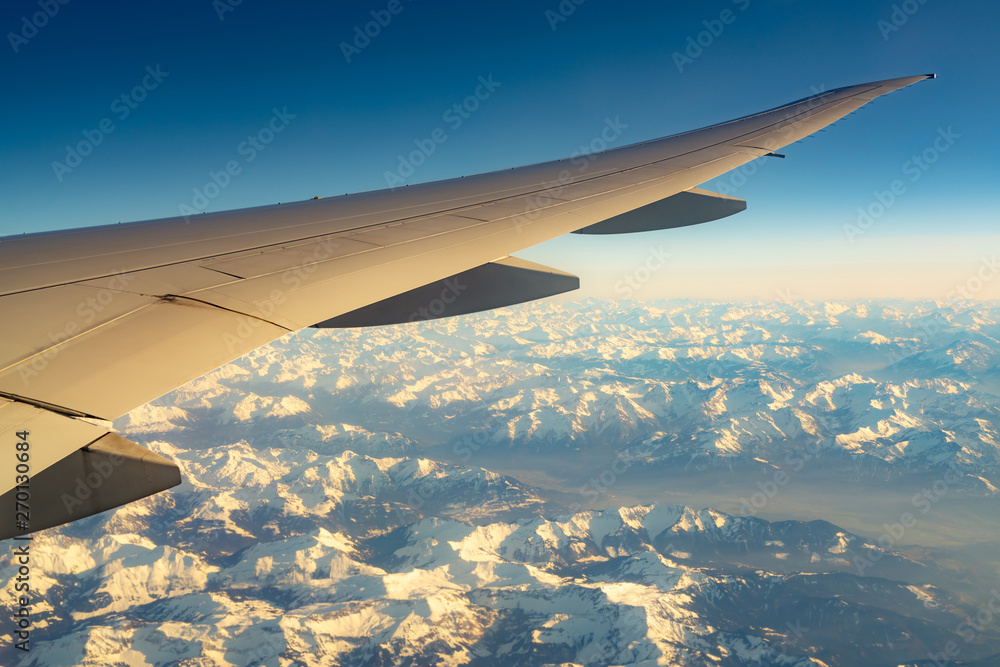 Wing of plane over mountain cover with white snow. Airplane flying on blue sky. Scenic view from airplane window. Commercial airline flight. Plane wing. Flight mechanics concept. International flight. Wing of plane over mountain cover with white snow. Airplane flying on blue sky. Scenic view from airplane window. Commercial airline flight. Plane wing. Flight mechanics concept. International flight.