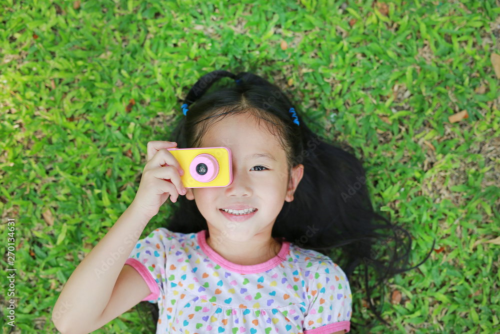 Cheerful little Asian girl takes photo with colorful digital camera lying on green lawn background. Cheerful little Asian girl takes photo with colorful digital camera lying on green lawn background.