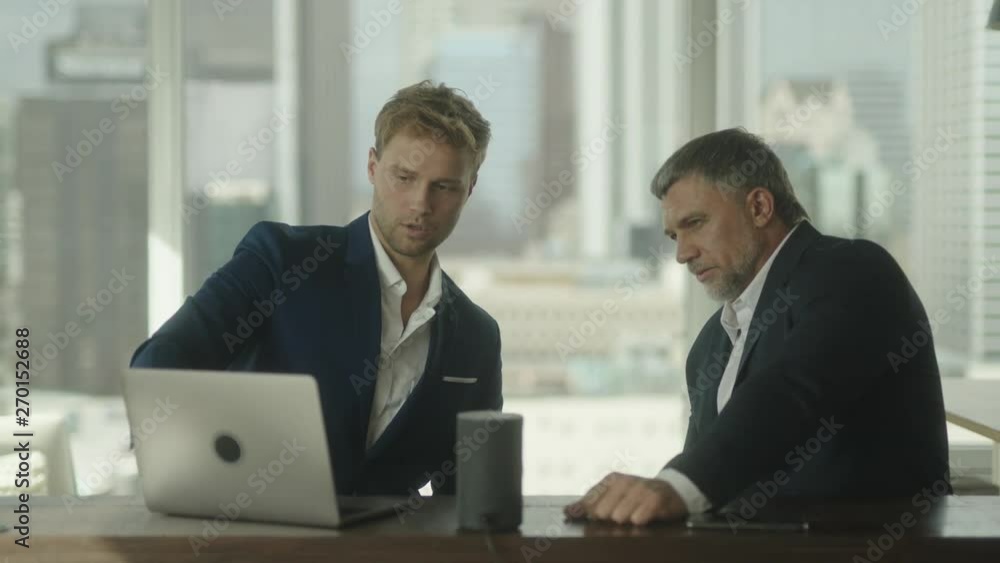 Two businessmen discuss project details that are showed on laptop in office with view on downtown. Two businessmen discuss project details that are showed on laptop in office with view on downtown.