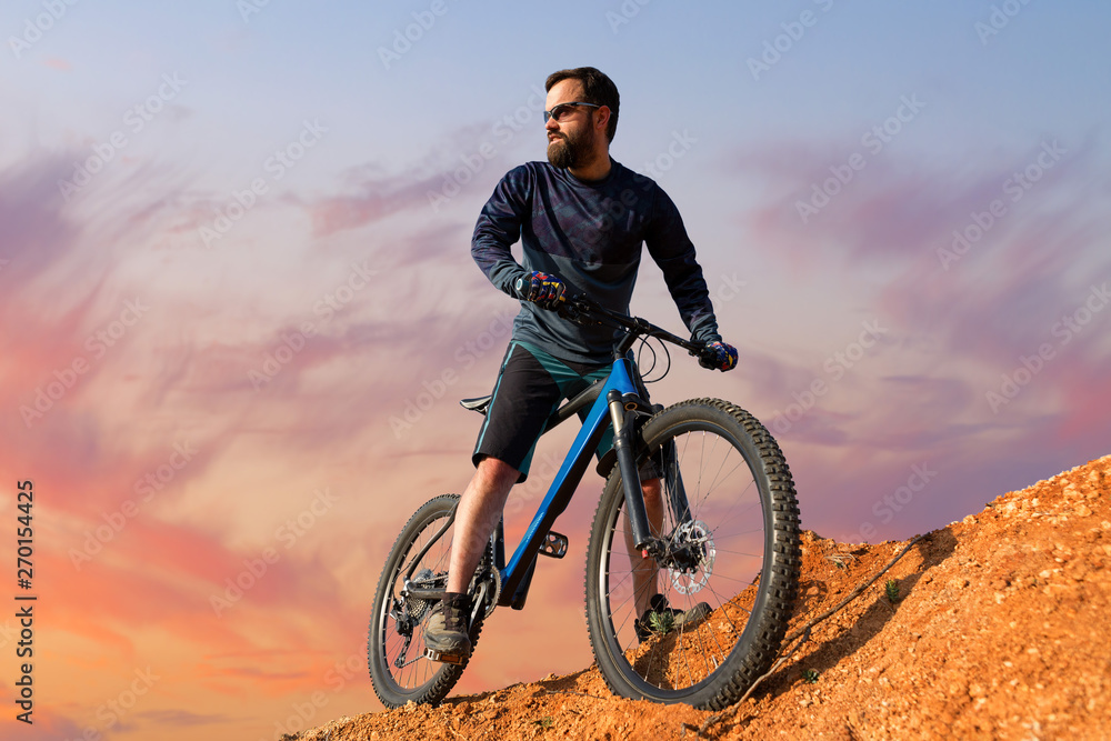 Naklejka premium Cyclist in shorts and jersey on a modern carbon hardtail bike with an air suspension fork rides off-road on the orange-red hills at sunset evening in summer