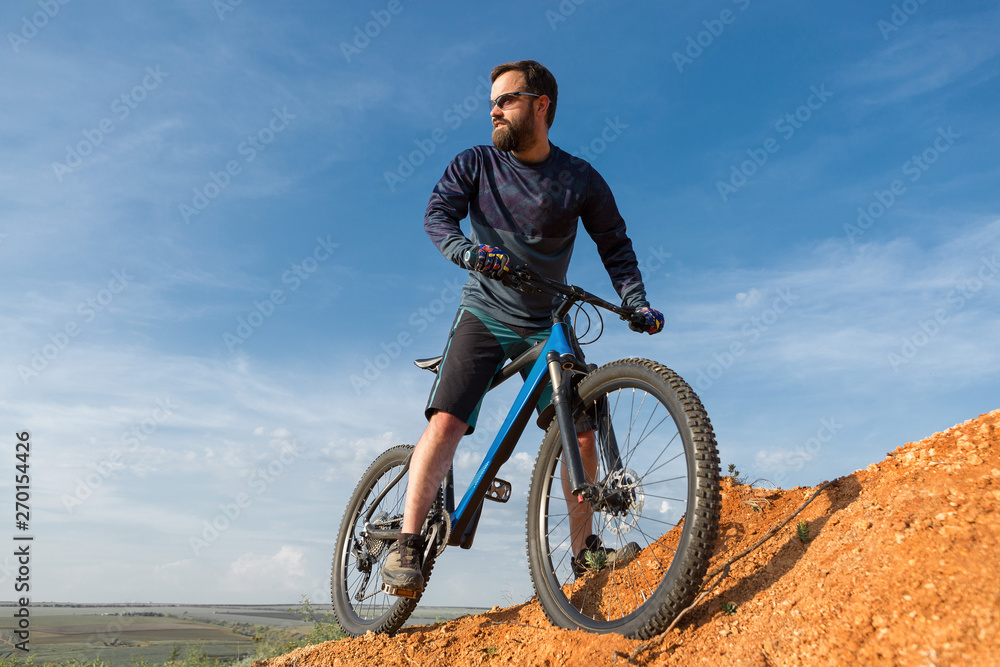 Fototapeta premium Rowerzysta w szortach i koszulce na nowoczesnym rowerze typu hardtail z włókna węglowego z widelcem pneumatycznym jeździ w terenie po pomarańczowo-czerwonych wzgórzach o zachodzie słońca w lecie