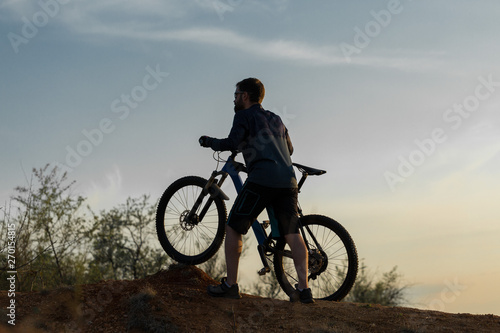 Fototapeta Naklejka Na Ścianę i Meble -  Cyclist in shorts and jersey on a modern carbon hardtail bike with an air suspension fork rides off-road on the orange-red hills at sunset evening in summer