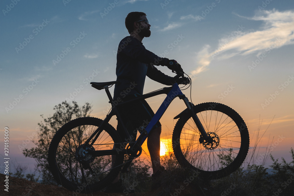 Obraz premium Cyclist in shorts and jersey on a modern carbon hardtail bike with an air suspension fork rides off-road on the orange-red hills at sunset evening in summer