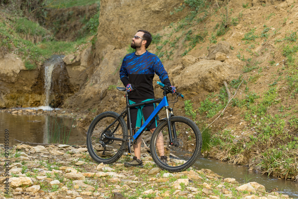 Cyclist in shorts and jersey on a modern carbon hardtail bike with an air suspension fork stands on the rocky shore of a mountain river Cyclist in shorts and jersey on a modern carbon hardtail bike with an air suspension fork stands on the rocky shore of a mountain river