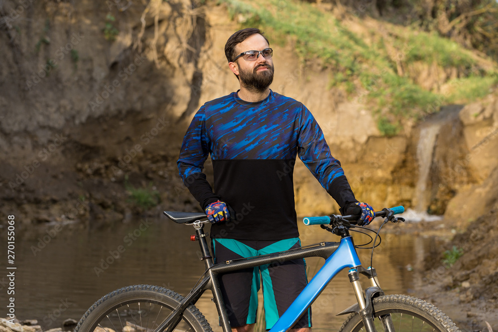Obraz premium Cyclist in shorts and jersey on a modern carbon hardtail bike with an air suspension fork stands on the rocky shore of a mountain river