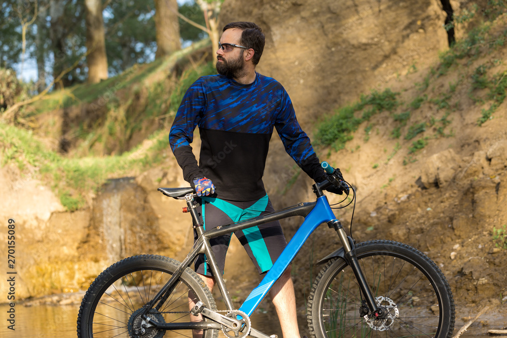 Fototapeta premium Cyclist in shorts and jersey on a modern carbon hardtail bike with an air suspension fork stands on the rocky shore of a mountain river