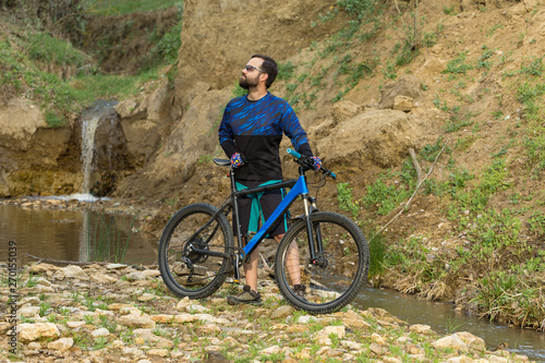 Fototapeta Naklejka Na Ścianę i Meble -  Cyclist in shorts and jersey on a modern carbon hardtail bike with an air suspension fork stands on the rocky shore of a mountain river