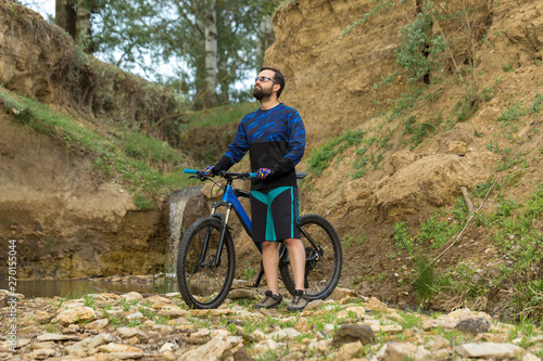 Cyclist in shorts and jersey on a modern carbon hardtail bike with an air suspension fork stands on the rocky shore of a mountain river