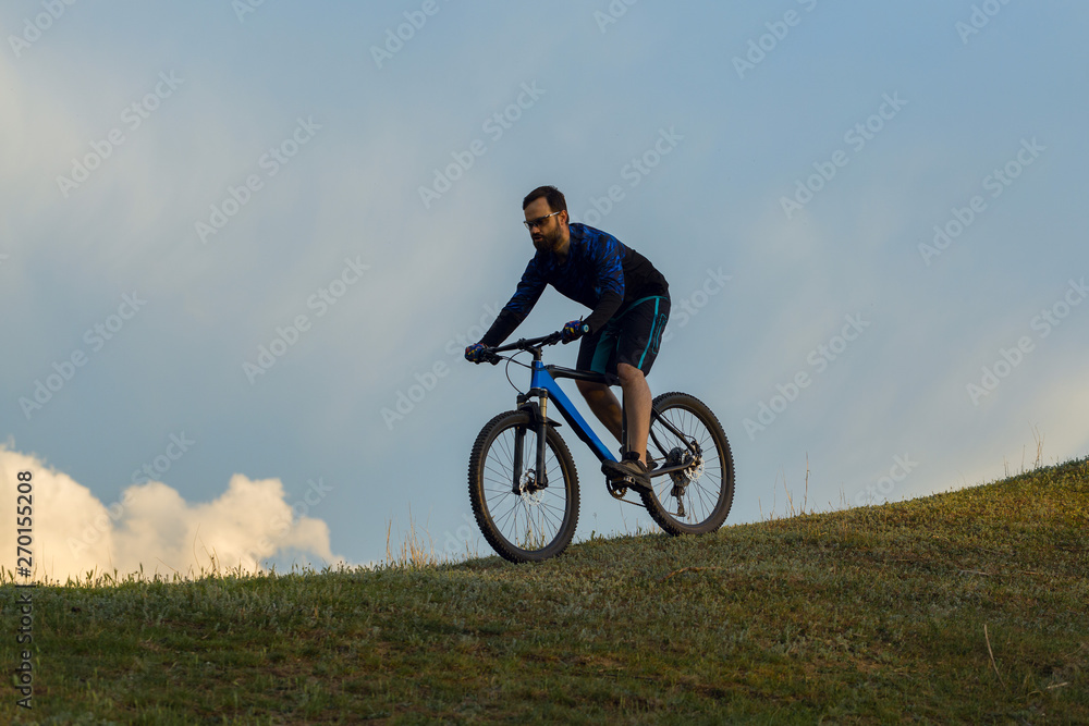 Cyclist in shorts and jersey on a modern carbon hardtail bike with an air suspension fork rides off-road on green hills near the forest	