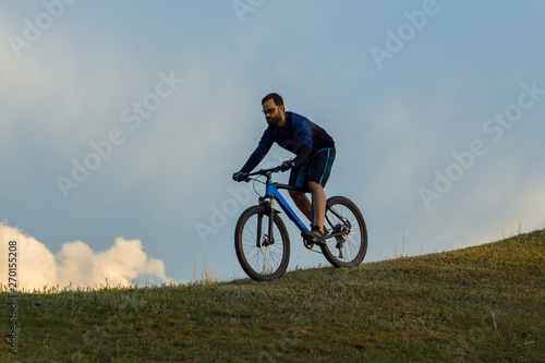 Cyclist in shorts and jersey on a modern carbon hardtail bike with an air suspension fork rides off-road on green hills near the forest	