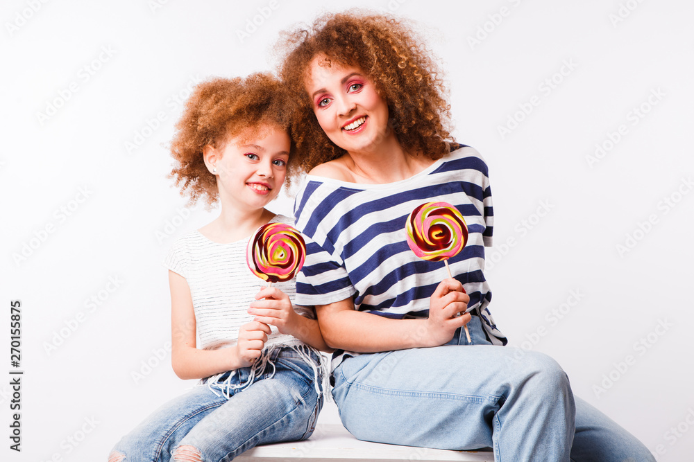 Mom and daughter with natural small curls on the head with bright lollipops, on a light background. Space for text. Mom and daughter with natural small curls on the head with bright lollipops, on a light background. Space for text.