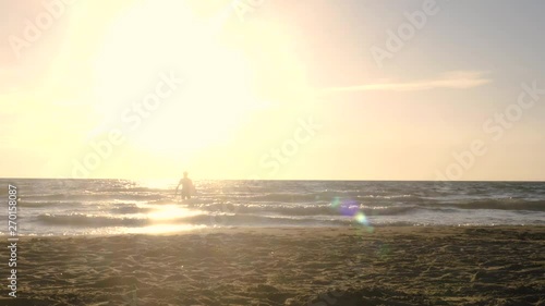 Young man surfer in wetsuit picks up surf board lying down on the sand at the beach and walks into the calm ocean water at sunset