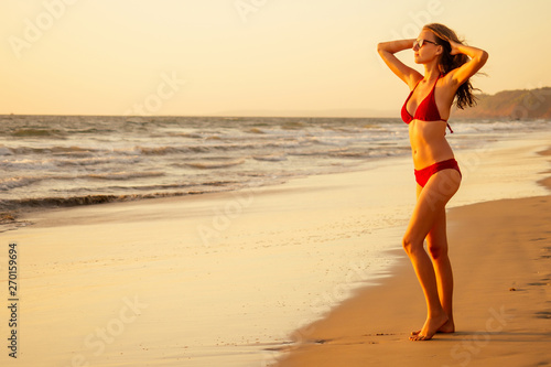 Beautiful sexy tanned model in red colour swimsuit on sand beach near ocean at sunset. Travel concept spf sunscreen sun protect