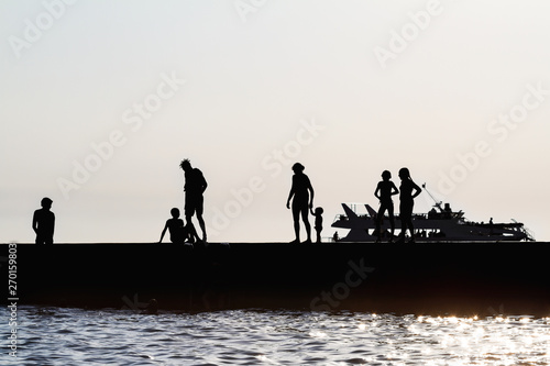 Silhouettes of people having a rest on a pier on the seashore. Rest on the sea beach with the boat on the background. © marketlan