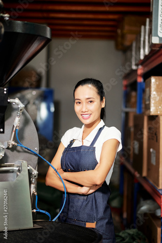 Portrait of Asian young female worker in apron standing with arms crossed and smiling at camera with machinery equipment in the background