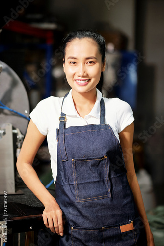 Portrait of Asian young female master standing in apron and smiling at camera while working on the factory