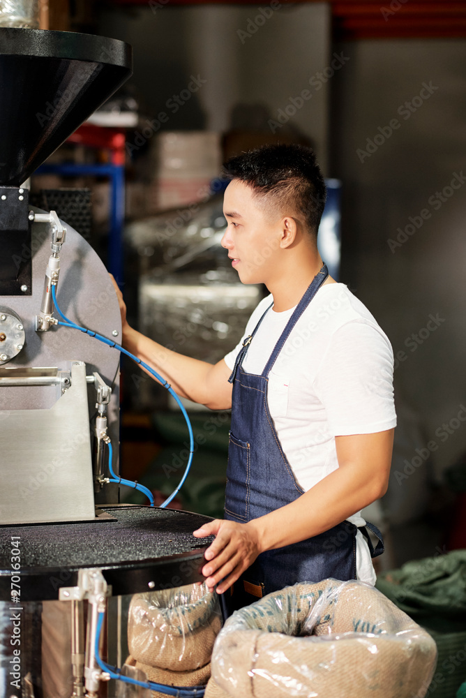 Young male factory worker in apron standing near the machine and controlling the process of production on the plant Young male factory worker in apron standing near the machine and controlling the process of production on the plant
