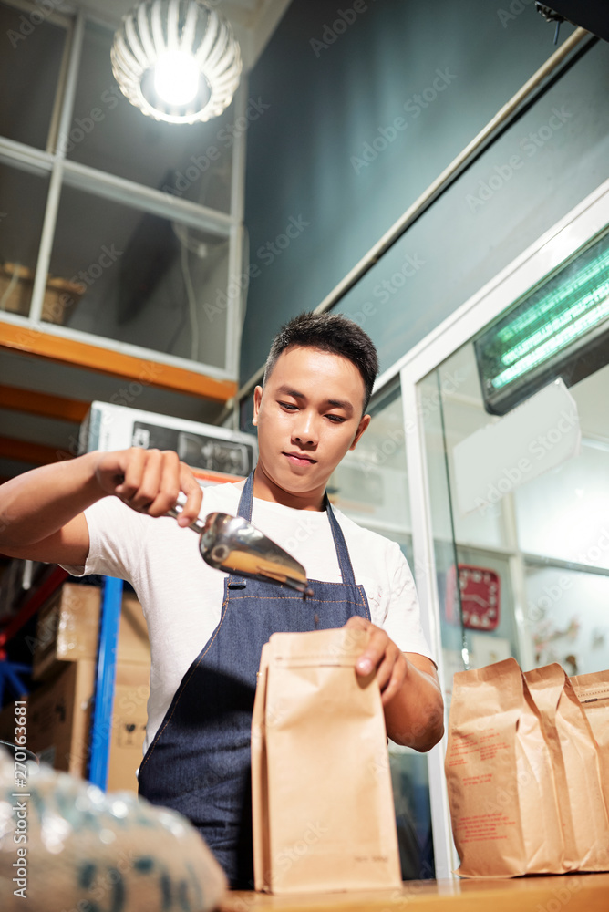 Asian young man standing in apron and filling paper bag with roasted coffee beans for sale after its production Asian young man standing in apron and filling paper bag with roasted coffee beans for sale after its production