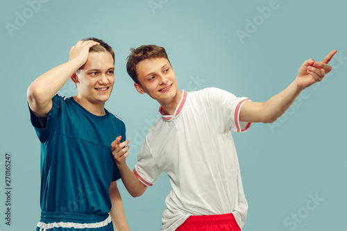 Two young men standing in sportwear isolated on blue studio background. Fans of sport, football or soccer club or team. Friends' half-length portrait. Concept of human emotions, facial expression.