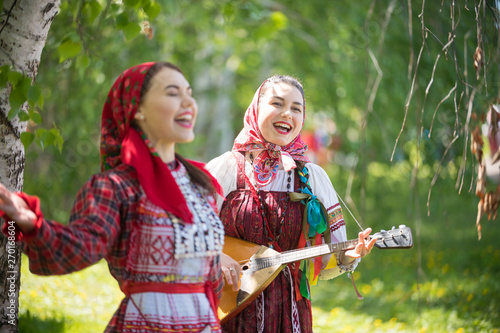 Two young attractive women in traditional russian clothes singing in the forest. One of them playing balalaika photo