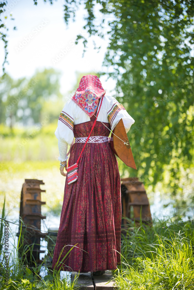Young woman in traditional russian clothes stands on a small bridge near the lake, with a balalaika behind her back