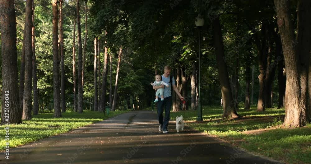 Young caucasian father walking in park with baby child on hands and white westie dog near during the sunset trees and grass Young caucasian father walking in park with baby child on hands and white westie dog near during the sunset trees and grass