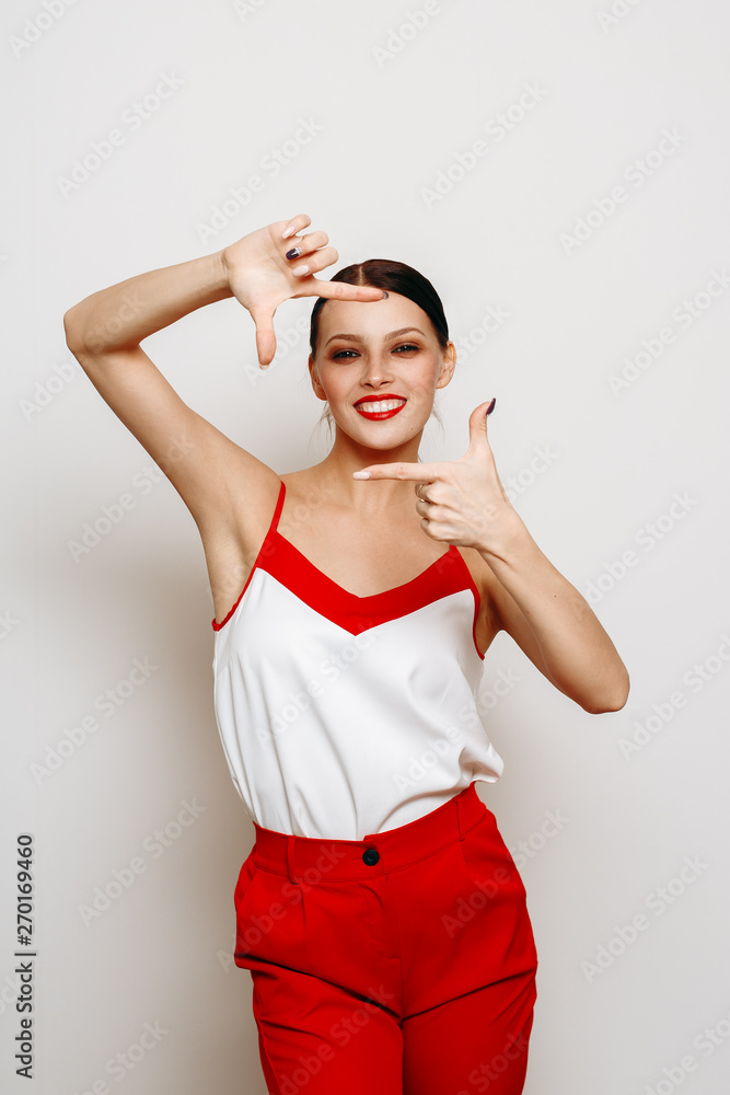 Human emotion. Beautiful brunette girl in a white and red top posing in the Studio on a white background. Woman positive smiling and looking at the camera, through the frame of her hands Human emotion. Beautiful brunette girl in a white and red top posing in the Studio on a white background. Woman positive smiling and looking at the camera, through the frame of her hands