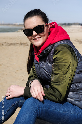 Young woman on the beach in warm clothes. A girl in a red hoodie wears a hood over her head. © evelinphoto
