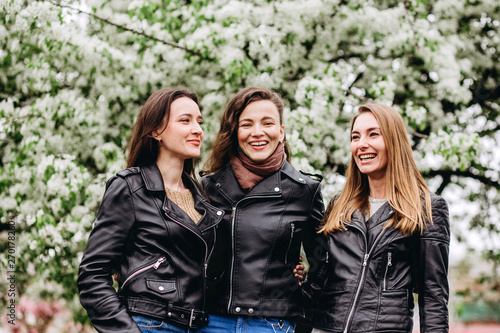 happy friends posing near blooming sakura flowers at cherry blossom trees during spring. Three young girls in leather jackets. Cute girl walks on nature.