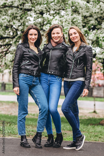 happy friends posing near blooming sakura flowers at cherry blossom trees during spring. Three young girls in leather jackets. Cute girl walks on nature.