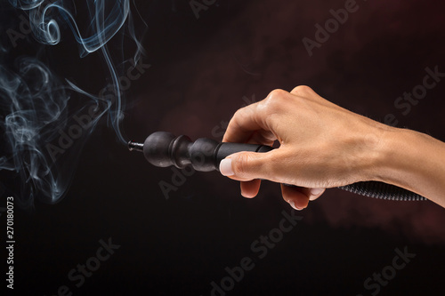 Hookah mouthpiece with a stream of smoke in a beautiful female hand on a dark background. Copy space.