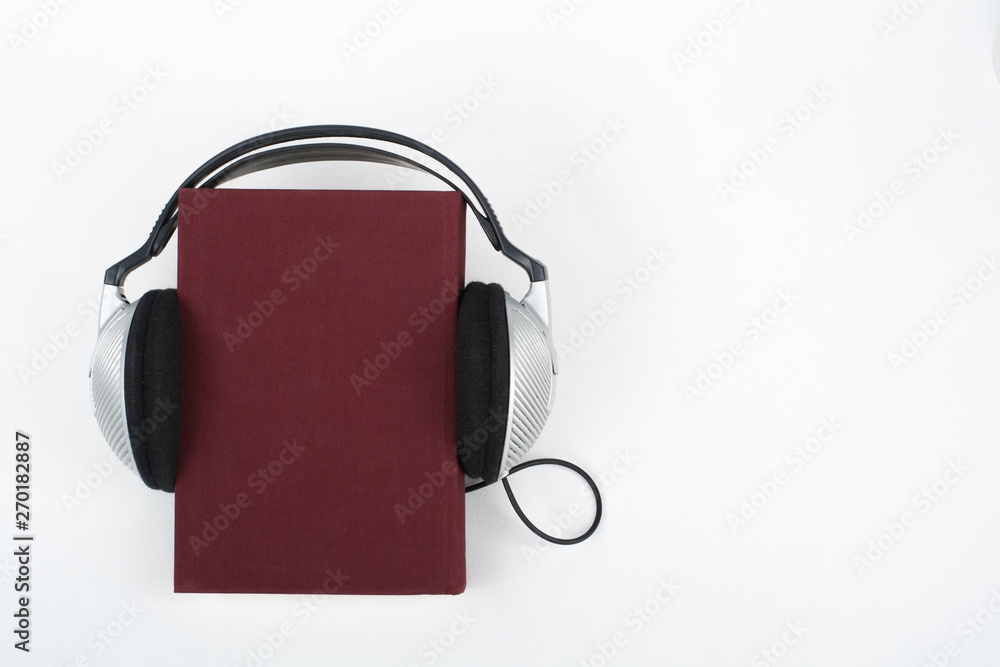 Audiobook on white background. Headphones put over red hardback book, empty cover, copy space for ad text. Distance education, e-learning concept. Top view Audiobook on white background. Headphones put over red hardback book, empty cover, copy space for ad text. Distance education, e-learning concept. Top view