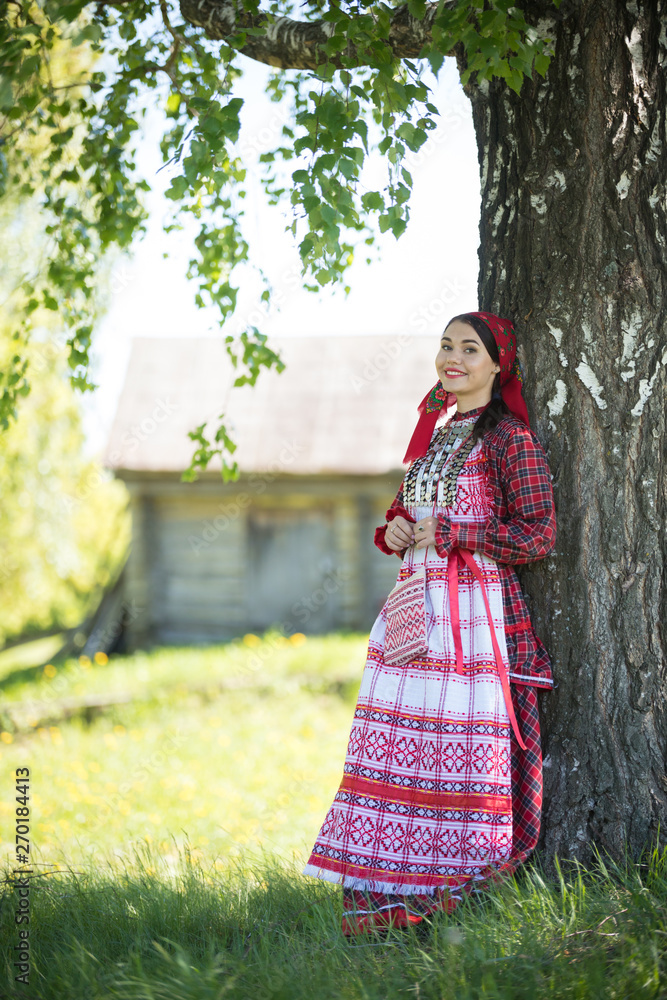 Young woman in traditional russian clothes standing under a tree and posing for a photo
