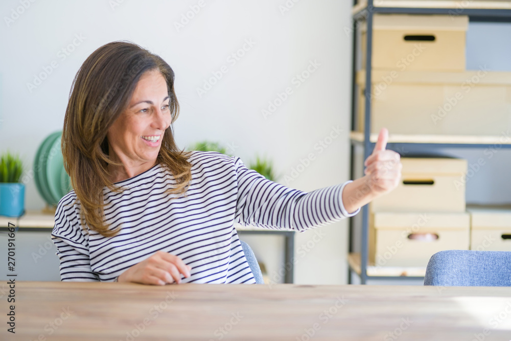 Middle age senior woman sitting at the table at home Looking proud, smiling doing thumbs up gesture to the side Middle age senior woman sitting at the table at home Looking proud, smiling doing thumbs up gesture to the side