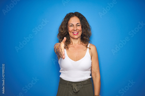 Middle age senior woman standing over blue isolated background smiling friendly offering handshake as greeting and welcoming. Successful business. © Krakenimages.com