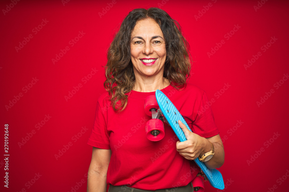 Middle age senior skater woman holding skateboard over red isolated background with a happy face standing and smiling with a confident smile showing teeth Middle age senior skater woman holding skateboard over red isolated background with a happy face standing and smiling with a confident smile showing teeth