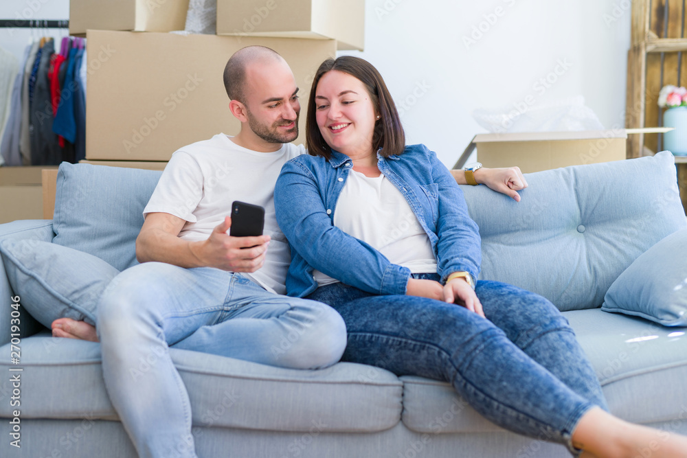Young couple sitting on the sofa at new home using smartphone, hugging and smiling happy for moving to a new apartment
