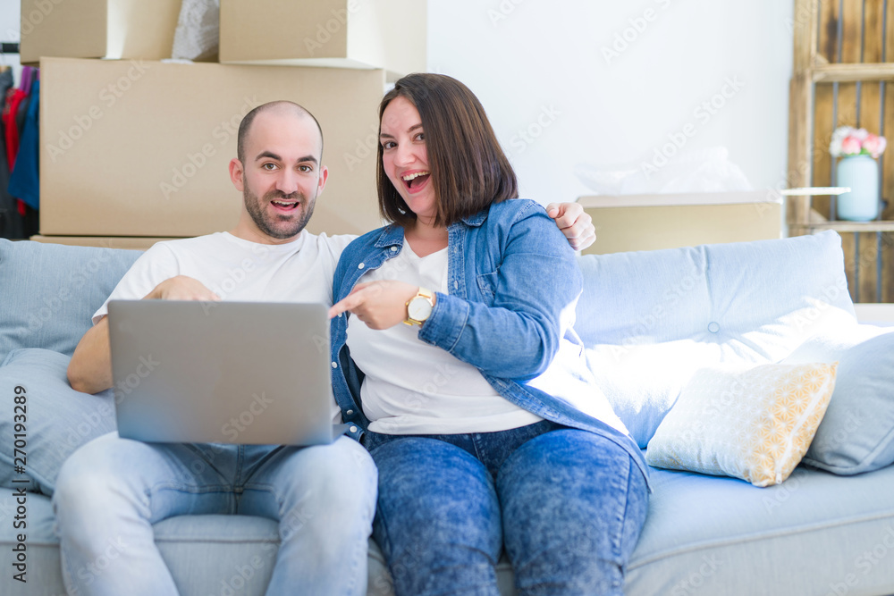 Young couple sitting on the sofa arround cardboard boxes moving to a new house using laptop very happy pointing with hand and finger Young couple sitting on the sofa arround cardboard boxes moving to a new house using laptop very happy pointing with hand and finger
