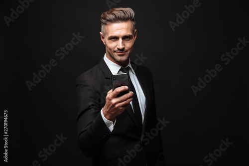 Business man posing isolated over black wall background using mobile phone. © Drobot Dean