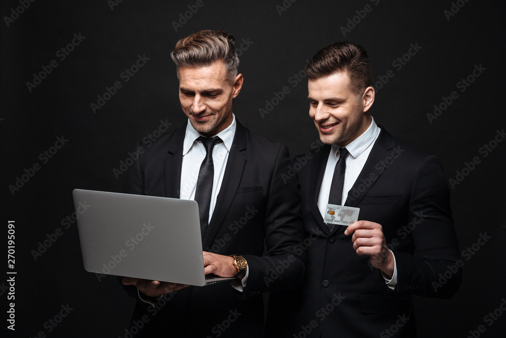 Handsome excited happy two business men posing isolated over black wall background using laptop computer holding credit card. Handsome excited happy two business men posing isolated over black wall background using laptop computer holding credit card.