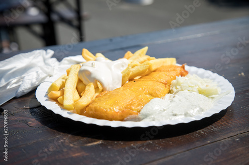 Seelachs Backfisch im Bierteig mit Remoulade, Pommes, Mayonnaise und Krautsalat draußen auf Pappteller und Holztisch 