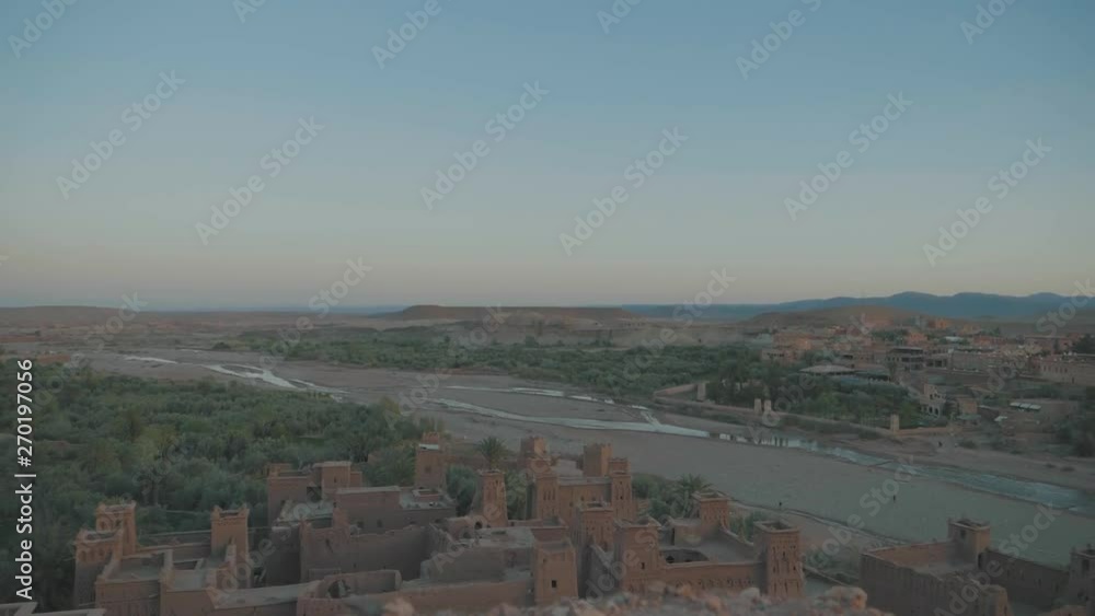 Standing on top a mountain overlooking an old Moroccan palace ruin at sunset with green shrubs and palm trees in the desert with orange sand and blue sky Standing on top a mountain overlooking an old Moroccan palace ruin at sunset with green shrubs and palm trees in the desert with orange sand and blue sky
